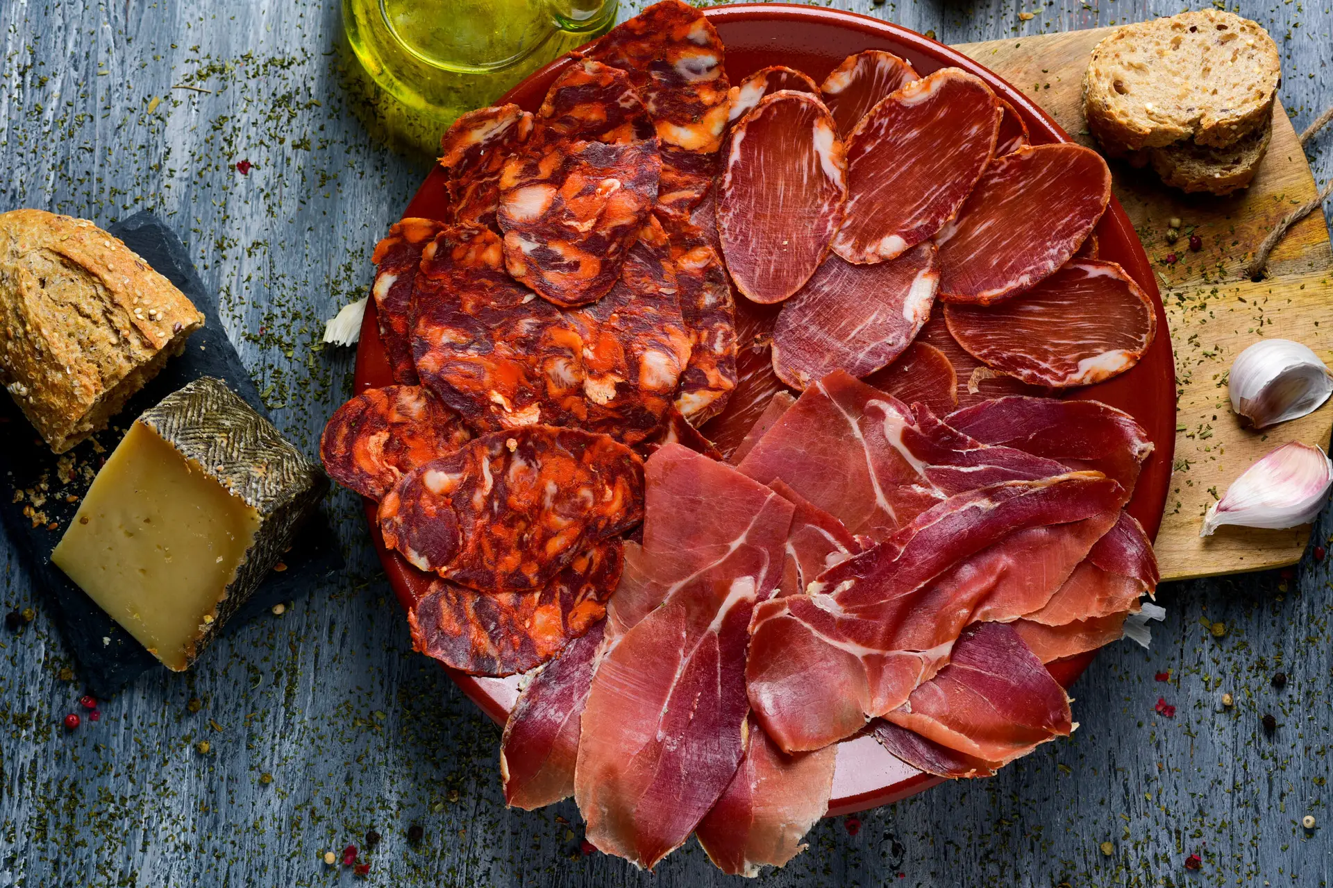 high-angle shot of a plate with an assortment of different spanish cold meats as chorizo, cured pork tenderloin and serrano ham, a piece of manchego cheese, bread and a cruet with olive oil on a table